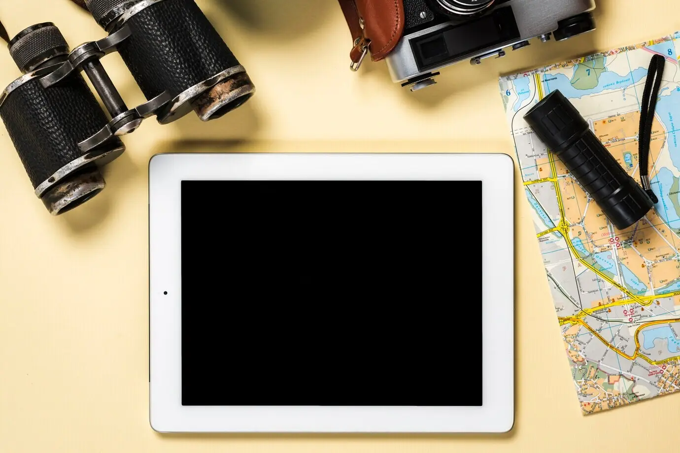 Fernglas, Kamera, Taschenlampe und Karte mit digitalem Tablet, das einen schwarzen Bildschirm anzeigt, vor beigem Hintergrund.