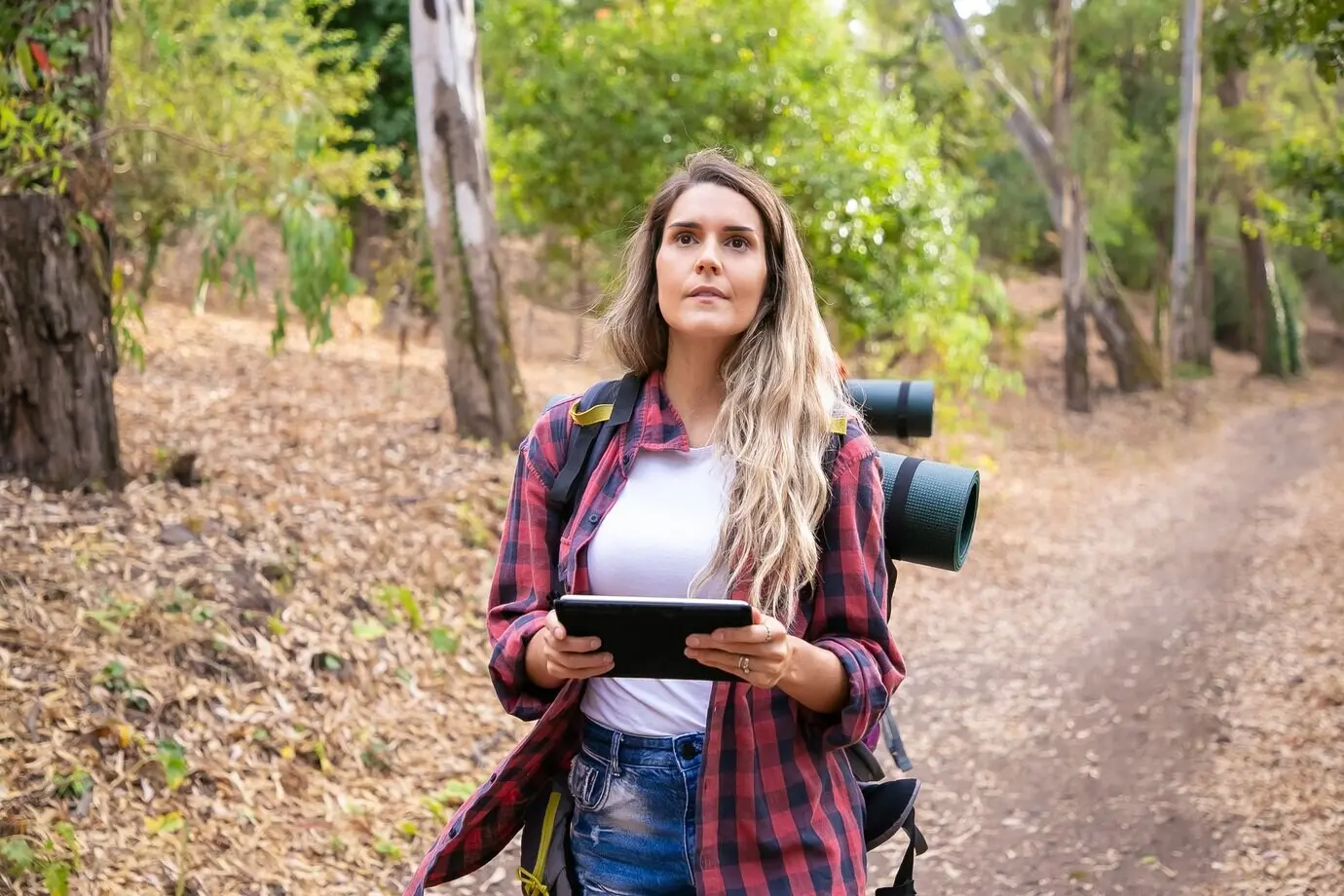 Hübsche Touristin hält ein Tablet mit einer Karte und blickt weg. Langhaarige kaukasische Frau wandert oder spaziert in der Natur und trägt Rucksäcke. Konzept für Backpacking-Tourismus, Abenteuer und Sommerurlaub.